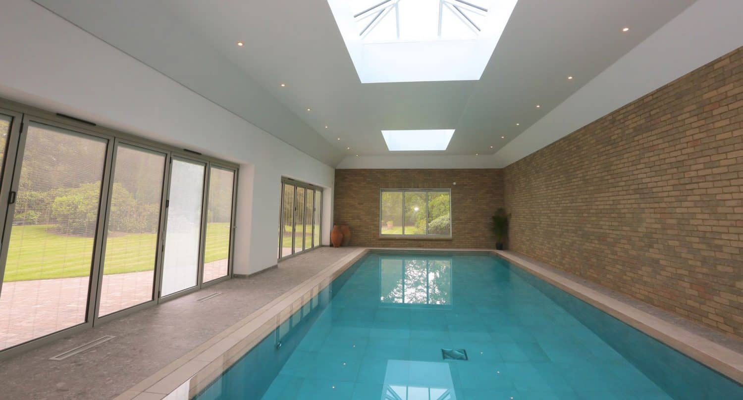 Indoor swimming pool with turquoise water, skylights above, brick wall on one side, and large glass doors and windows offering views of a green garden outside.