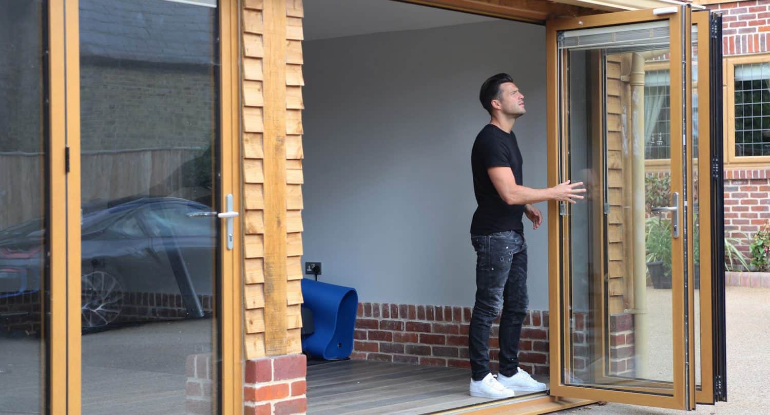 A man wearing a black t-shirt and jeans stands inside a modern room, sliding open a large glass folding door, with a car parked outside.