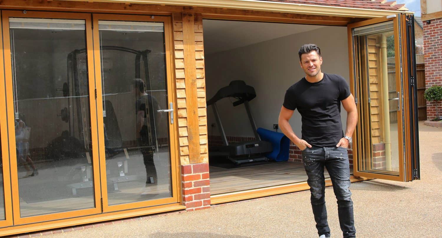 A man in a black t-shirt and jeans stands outside a modern home gym with large glass doors, with fitness equipment like a treadmill visible inside.