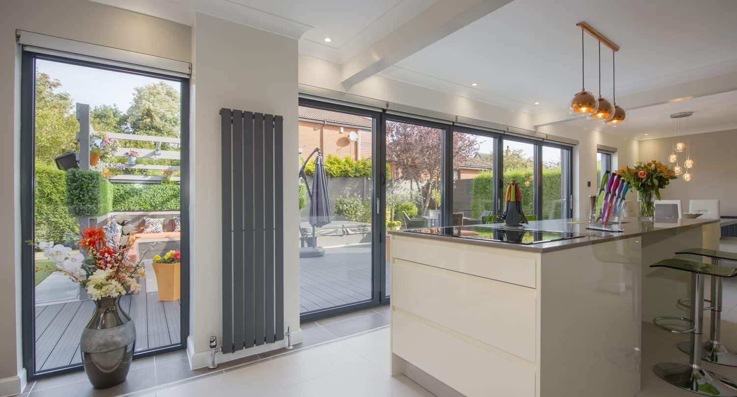 A modern kitchen with glossy white cabinets, an island with barstools, pendant lights, and large bifold doors opening to a patio and garden filled with greenery and flowers.