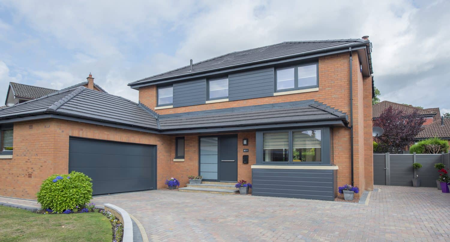 Modern two-story brick house with gray roof and accents, bespoke glass solutions such as sliding doors, attached garage, paved driveway, and a neatly landscaped yard with flowers and bushes.
