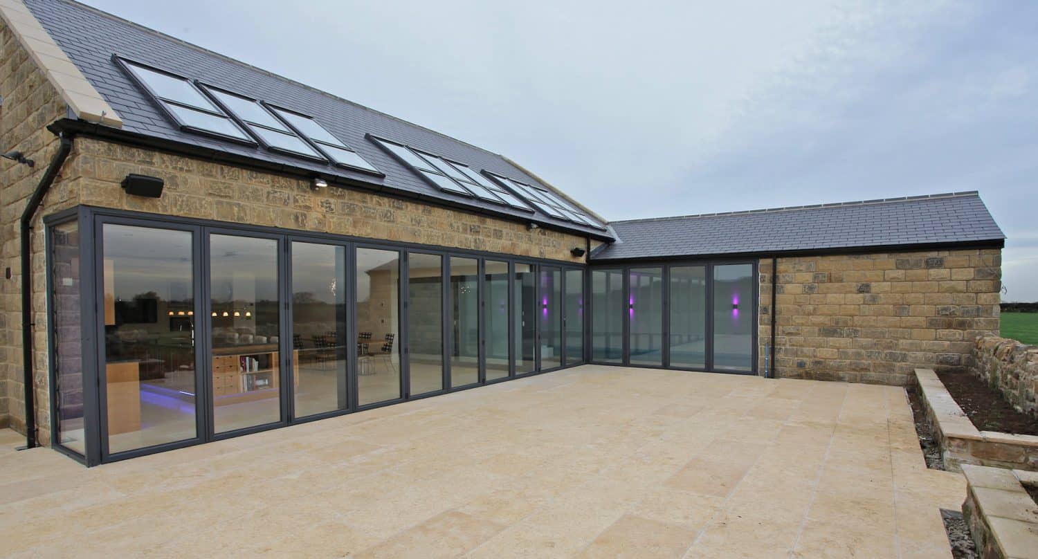 Modern stone house with large glass sliding doors and windows, opening onto a spacious tiled patio. The glass reflects the sky, and the house is surrounded by a low stone wall and open grassy area.