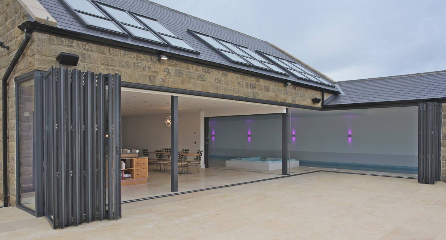 Modern house with large folding glass doors opened to reveal an indoor pool with purple accent lights, a spacious kitchen, and a dining area. The exterior features stone walls and a tiled roof with skylights.