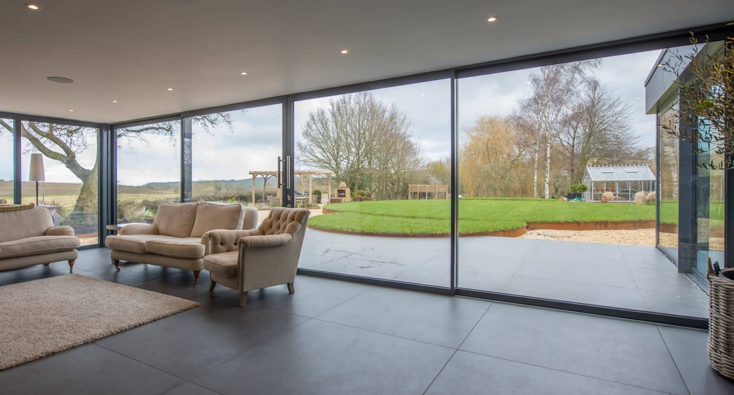 Modern living room with large floor-to-ceiling glass windows offering a clear view of a green lawn, trees, outdoor seating area, and a greenhouse on a cloudy day. Light-colored sofas and a rug are visible inside.