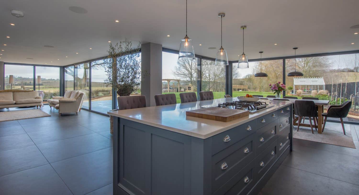Modern open-plan kitchen and dining area with large windows, a dark blue island with a stovetop, pendant lights, a wooden dining table, and views of a garden and countryside outside.
