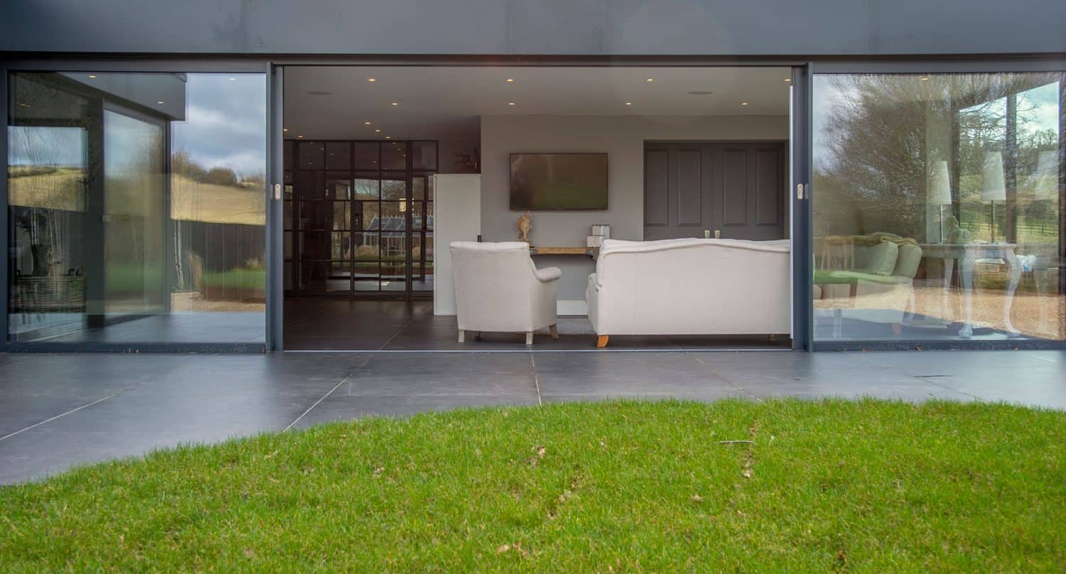 A modern living room with large glass sliding doors and bespoke glass solutions opens to a grassy yard, showing light-colored sofas, a TV on the wall, and recessed ceiling lights inside.