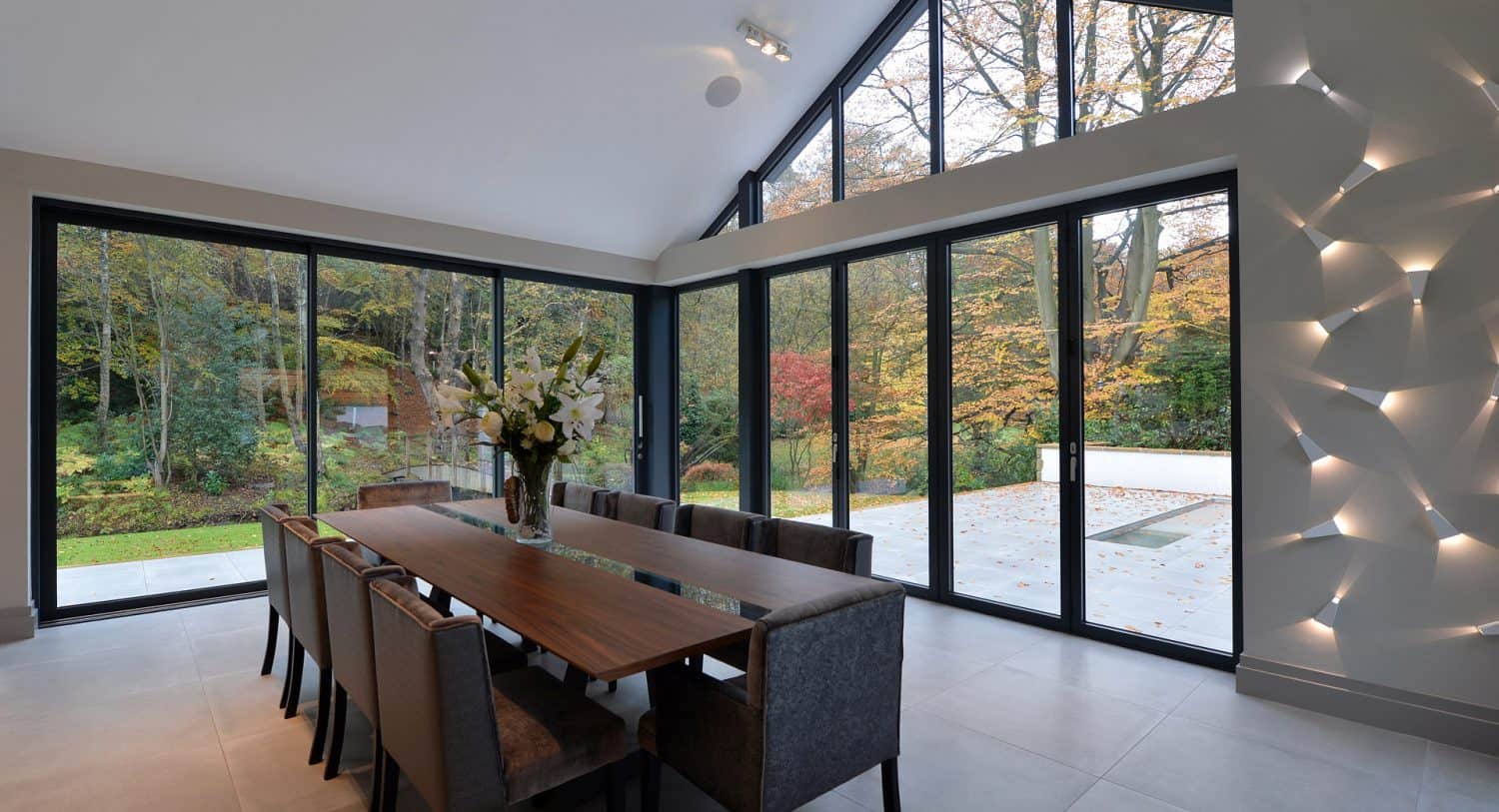 Modern dining room with a long wooden table, gray upholstered chairs, and large floor-to-ceiling windows overlooking a lush, leafy garden; wall with geometric lighting design.