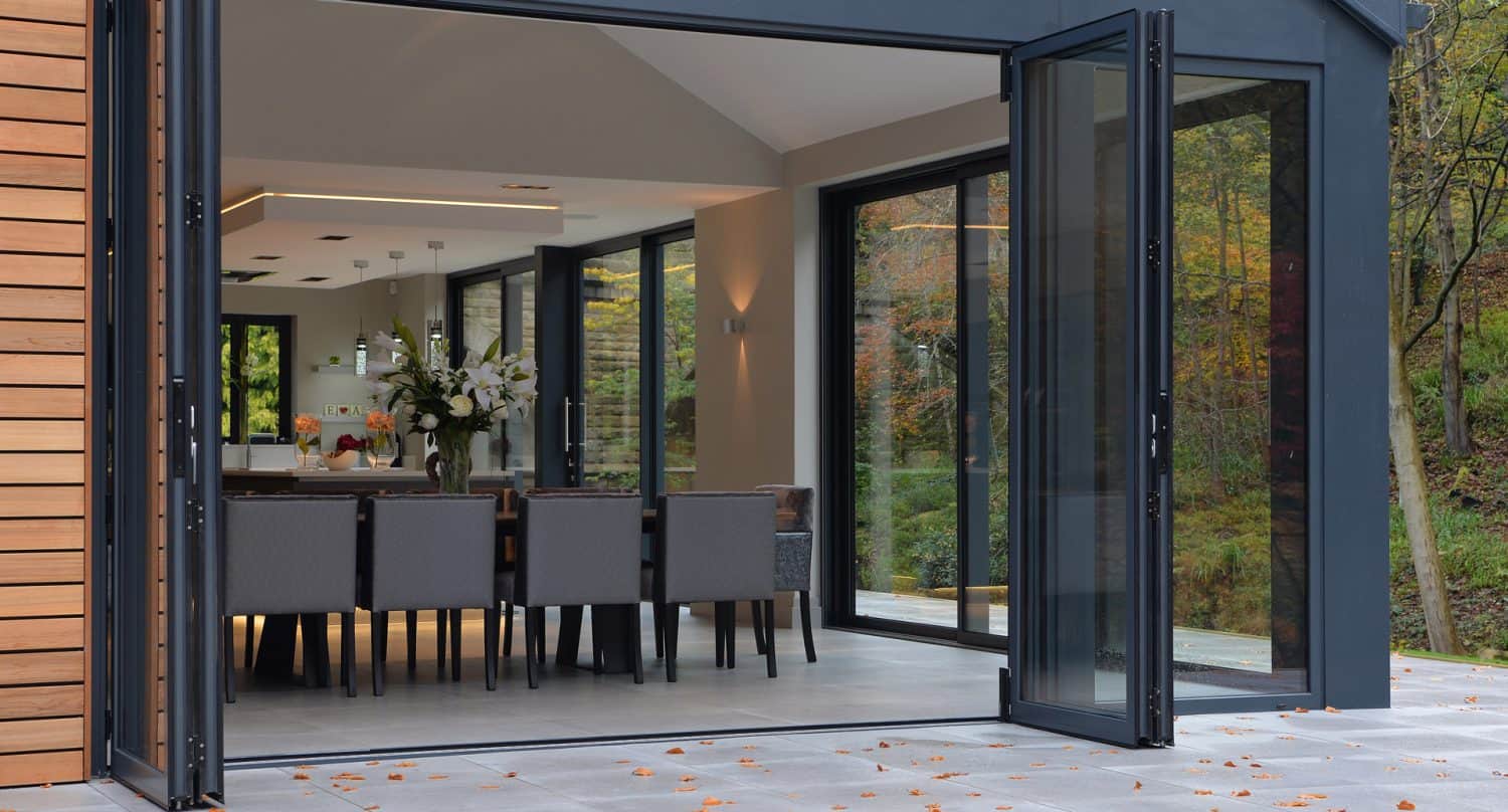 A modern dining room with folding glass doors fully opened, connecting the indoor space to the outside. The dining area features a long table with gray chairs, floral centerpiece, and views of trees outside.