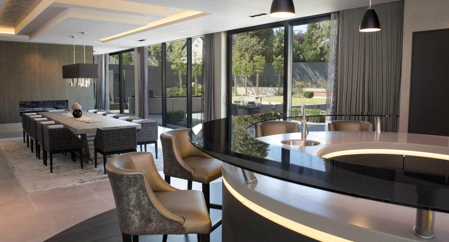 Modern open-plan dining and kitchen area with a long dining table, cushioned chairs, curved black countertop with bar stools, large windows, pendant lights, and views of a landscaped garden outside.