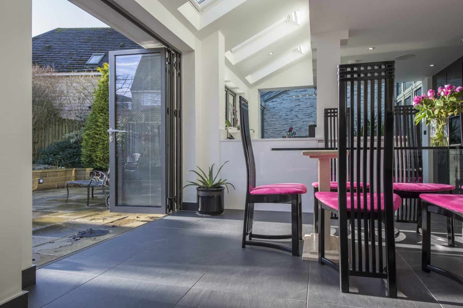 Modern dining area with black chairs featuring bright pink cushions, a glass table with a vase of flowers, and large sliding doors open to a sunny patio with plants and outdoor seating. Natural light fills the space.
