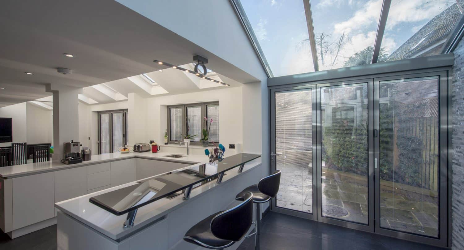 Modern kitchen with white cabinets, a sleek breakfast bar with two black stools, and large glass doors and ceiling letting in natural light, overlooking a patio area outside.