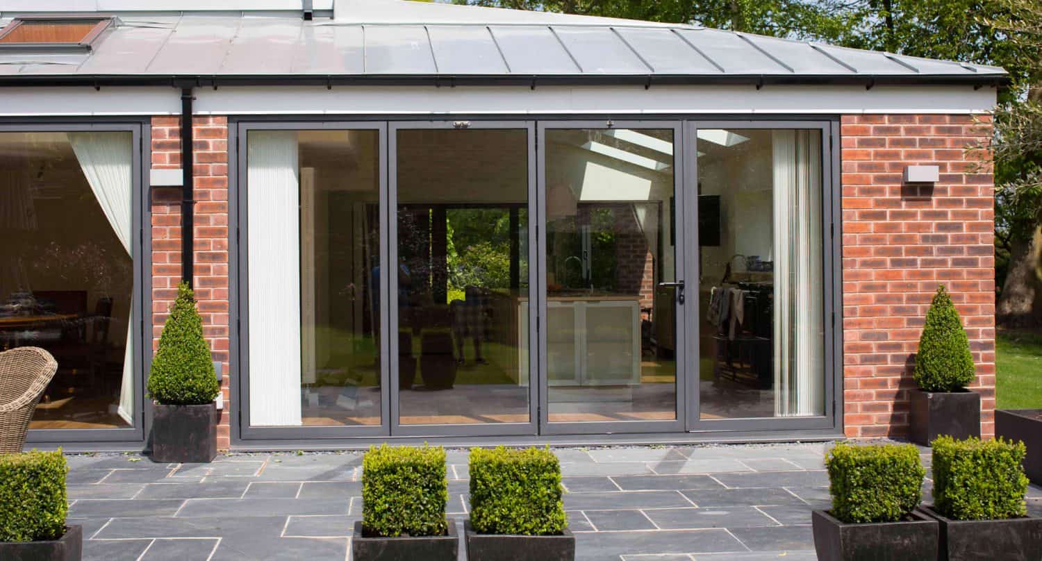 Modern brick house with large glass sliding doors, black frames, and a patio. Four square planters with trimmed green shrubs are placed along the tiled walkway in front of the doors.
