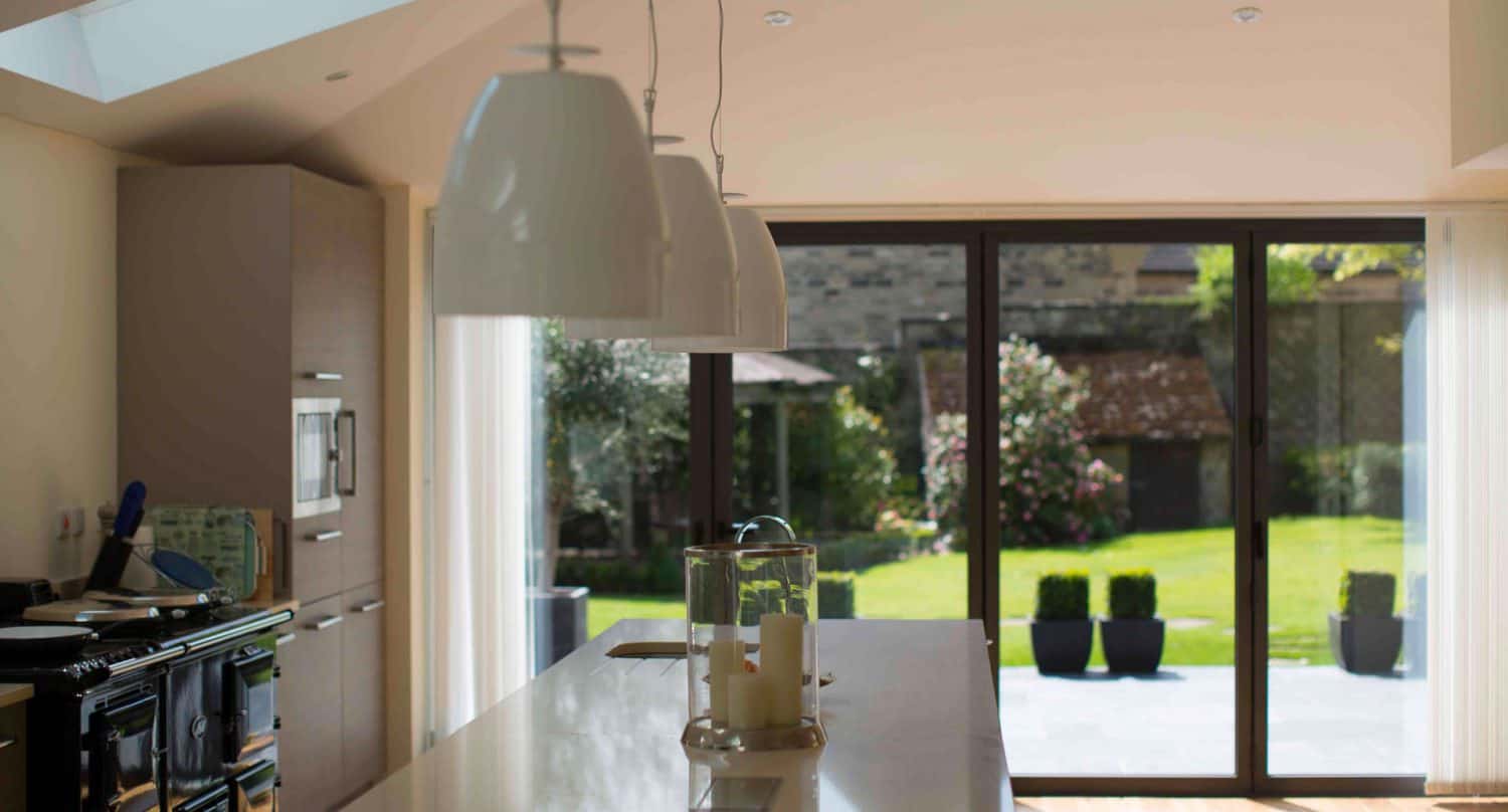 Modern kitchen with pendant lights over a white island, large glass doors opening to a sunny garden with green lawn, shrubs, and potted plants outside.