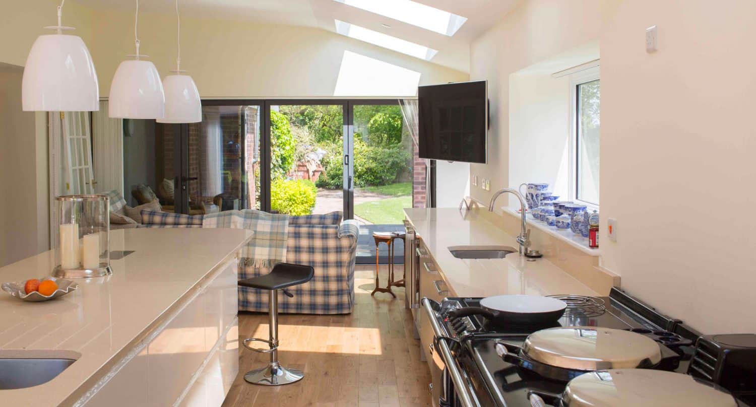 Bright, modern kitchen with glossy countertops, pendant lights, and a stove in the foreground. Large glass doors open to a green garden, and a checkered sofa sits in the adjacent living area.