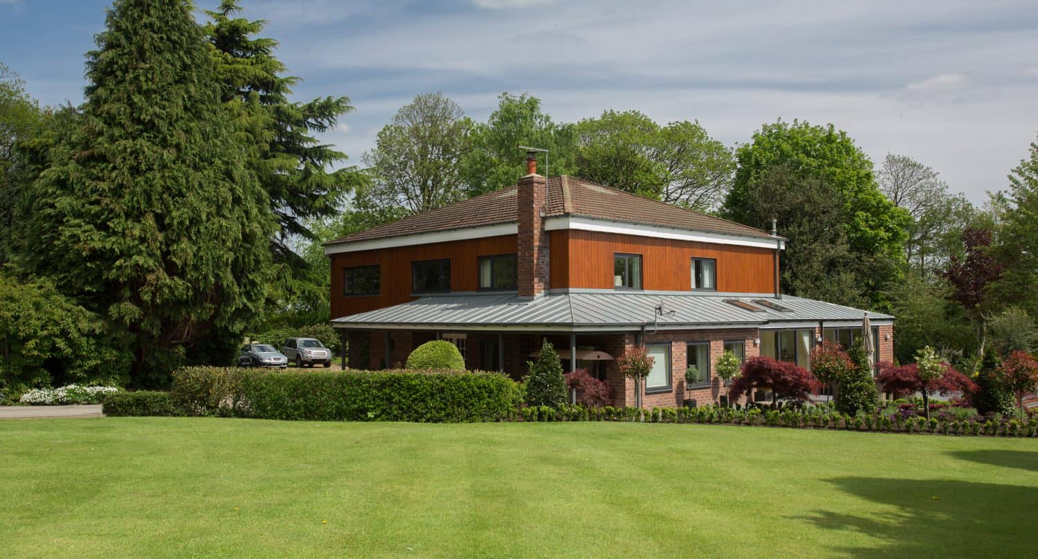 A modern two-story house with a mix of brick and wood exterior features bespoke glass solutions like sleek sliding doors, nestled among lush green trees, manicured lawns, and neatly trimmed hedges under a partly cloudy sky.