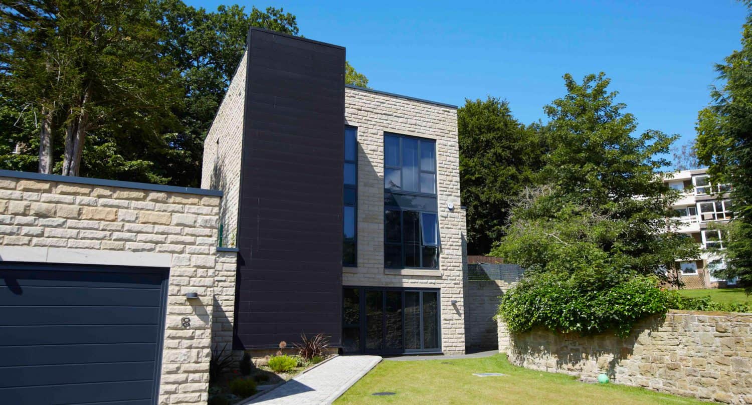 A modern two-story house with large windows, stone and dark panel exterior, and a neatly landscaped lawn, surrounded by trees and other buildings in the background.
