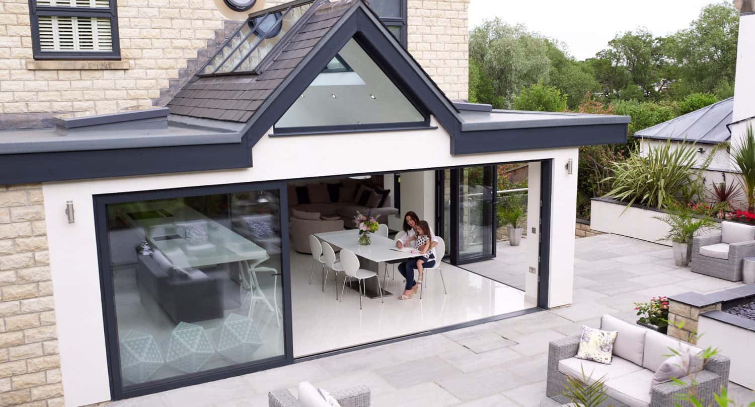 A modern house with large glass sliding doors opens to a patio. Inside, a woman sits at a dining table with a child, reading together. The kitchen and living area are visible; outdoor seating is arranged on the patio.