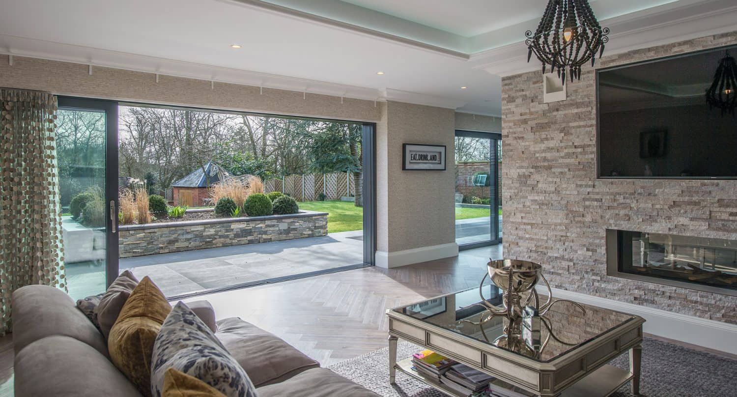 Modern living room with large sliding glass doors opening to a landscaped backyard. The room features a stone accent wall with a built-in fireplace, a stylish chandelier, and a mirrored coffee table.