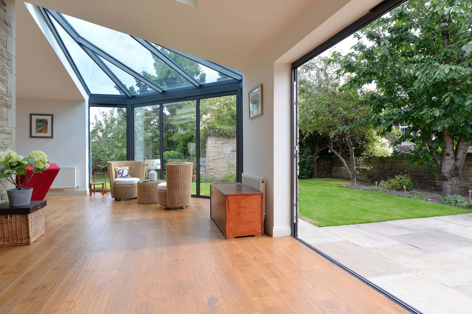 A bright sunroom with bespoke glass solutions features large glass windows and a glass roofing, wooden floor, wicker chairs, and open doors leading to a green garden with trees and a stone wall.