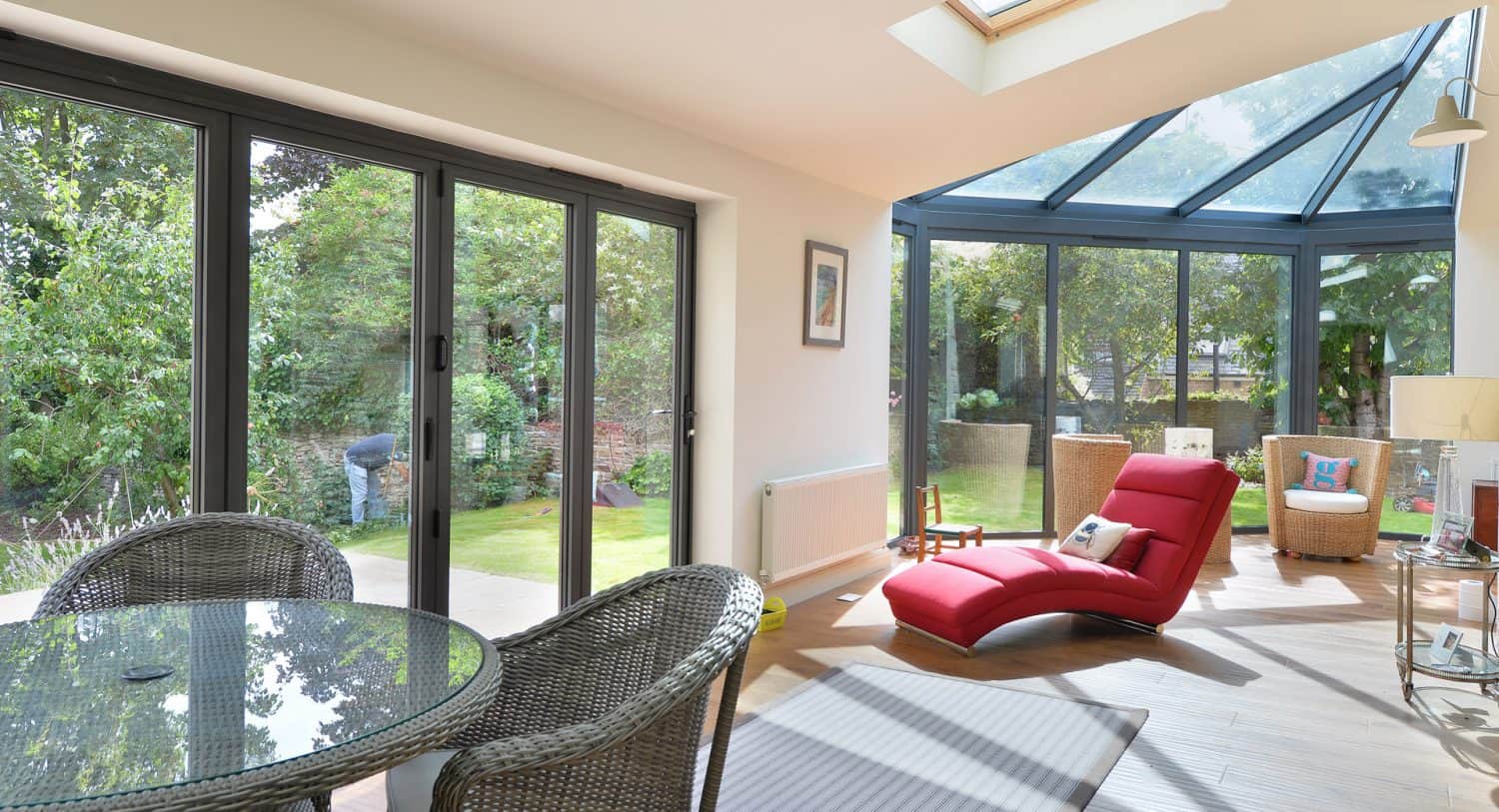 A bright sunroom with large glass windows and doors overlooking a garden. Inside, there’s a round glass table with wicker chairs and a red chaise lounge. Sunlight fills the space, creating a cozy and inviting atmosphere.