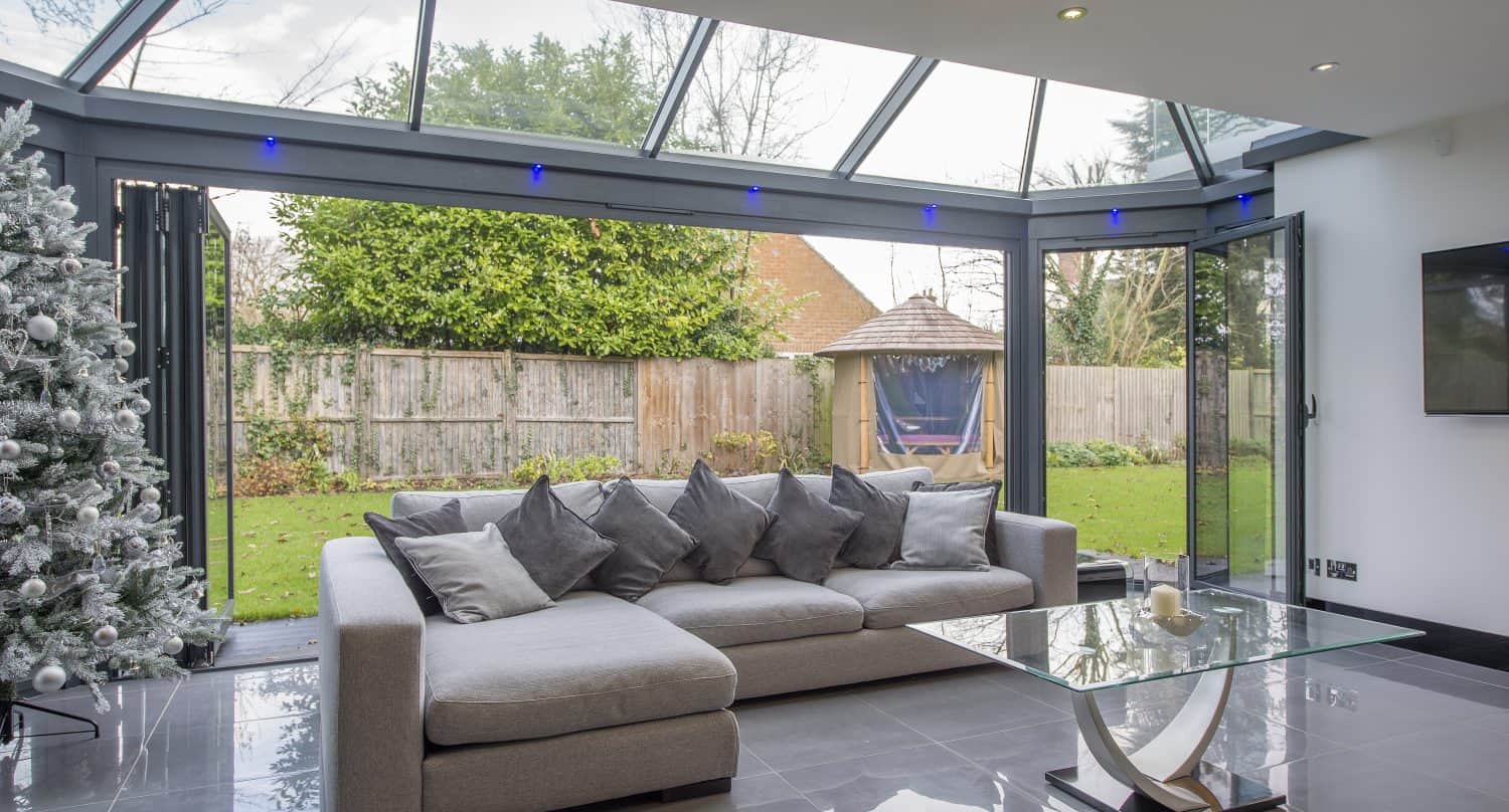 Modern living room with a grey sectional sofa, glass coffee table, and decorated Christmas tree, featuring floor-to-ceiling windows and open bifold doors looking out onto a garden with a wooden gazebo.