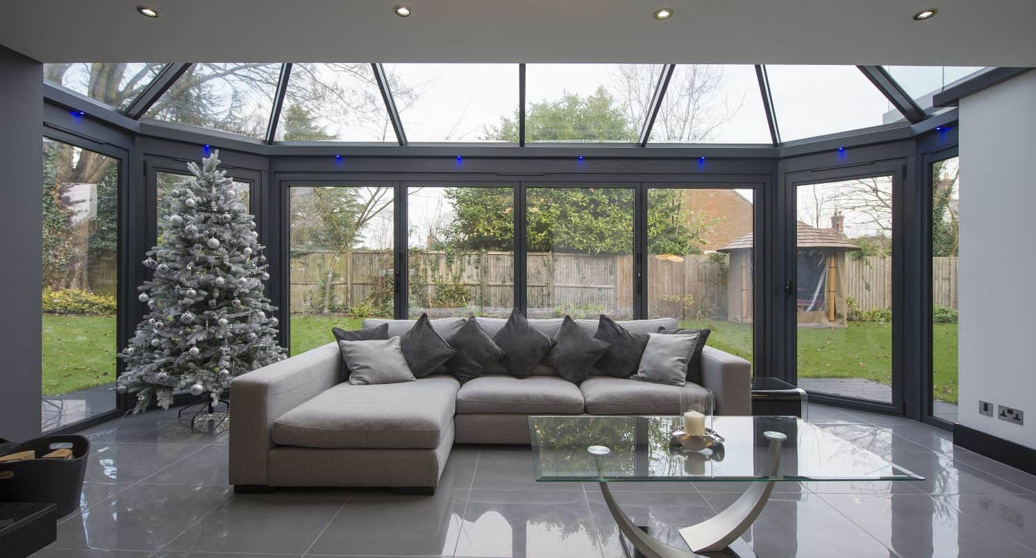 Modern living room with a large glass conservatory, grey corner sofa, glass coffee table, and a decorated white Christmas tree. Bifold doors and floor-to-ceiling windows overlook a garden with trees and grass.