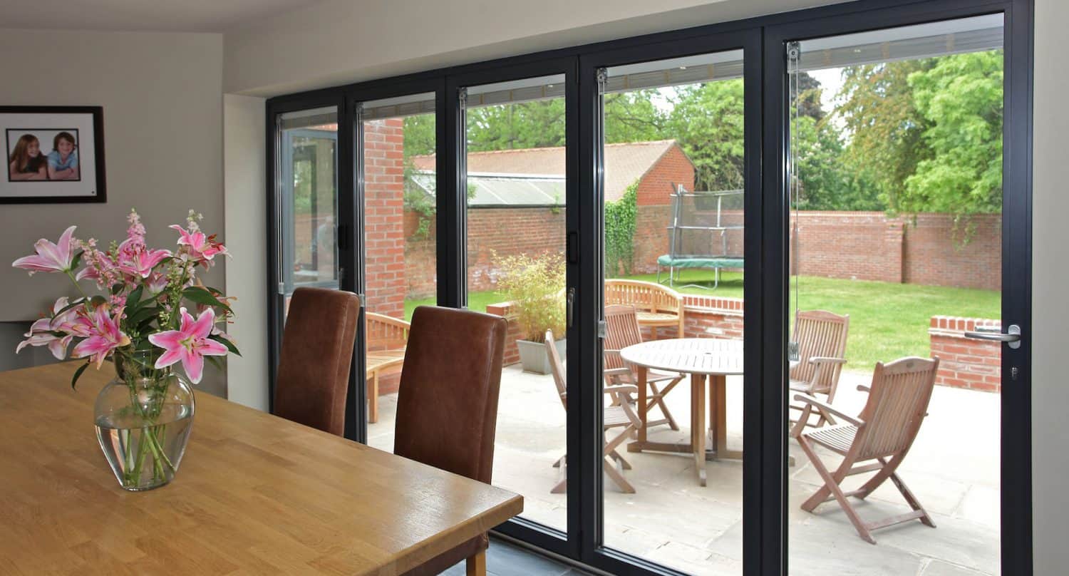 A bright dining area with a wooden table, pink flowers in a vase, brown chairs, and large glass folding doors that open to a patio with outdoor furniture, a trampoline, and a green garden.
