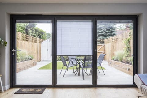 A view through large bifold doors reveals a modern patio with a table, four chairs, potted plants, a grassy area, wooden fences, and a small shed at the back—showcasing bespoke glass solutions for seamless indoor-outdoor living.