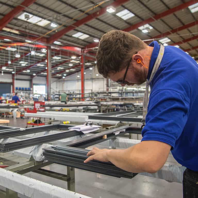 A man wearing ear protection and a blue shirt works with metal frames for bespoke glass solutions in a large, well-lit factory, surrounded by industrial equipment and high ceilings.