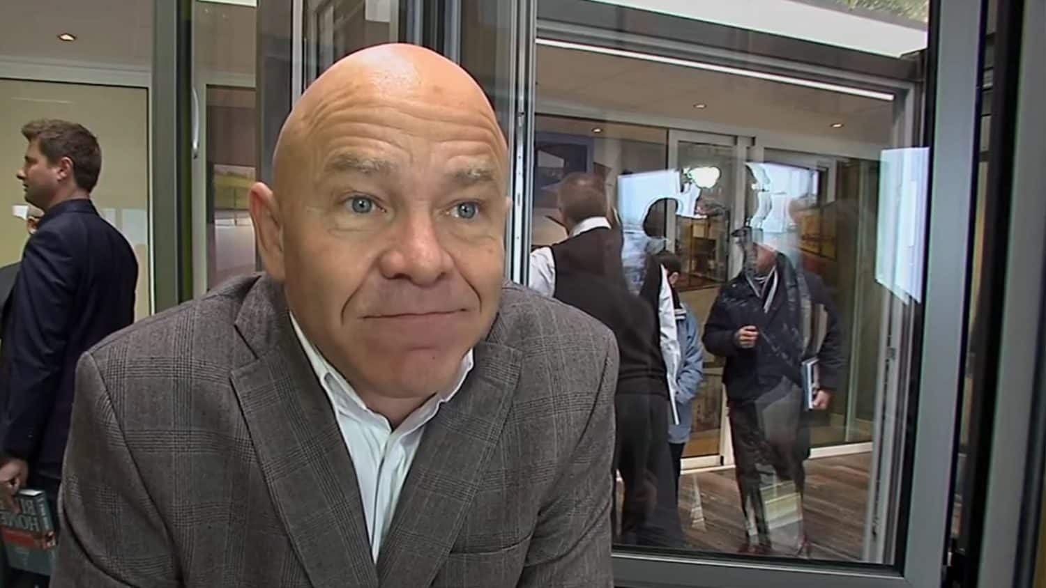 A bald man in a gray checked blazer looks into the camera with a slight smile. Several people are visible in the background, some walking through sliding doors beneath bright indoor lighting.