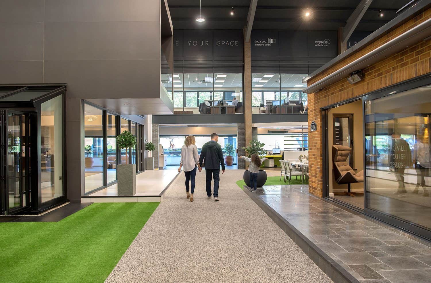 A man and woman walk hand-in-hand through a modern, open indoor space with artificial grass, large windows, and office areas. Sliding doors lead to the area, while another person sits on a bench nearby. The atmosphere is bright and contemporary.