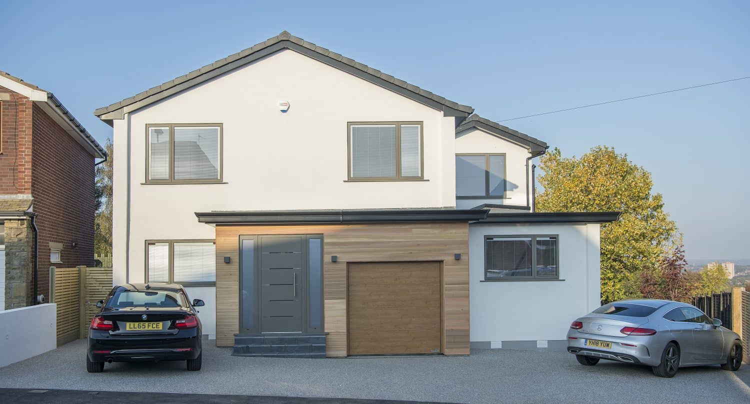 A modern two-story house with white walls and wood accents features a gray front door and bespoke glass solutions. Two cars are parked in the driveway, and trees are visible in the background under a clear sky.