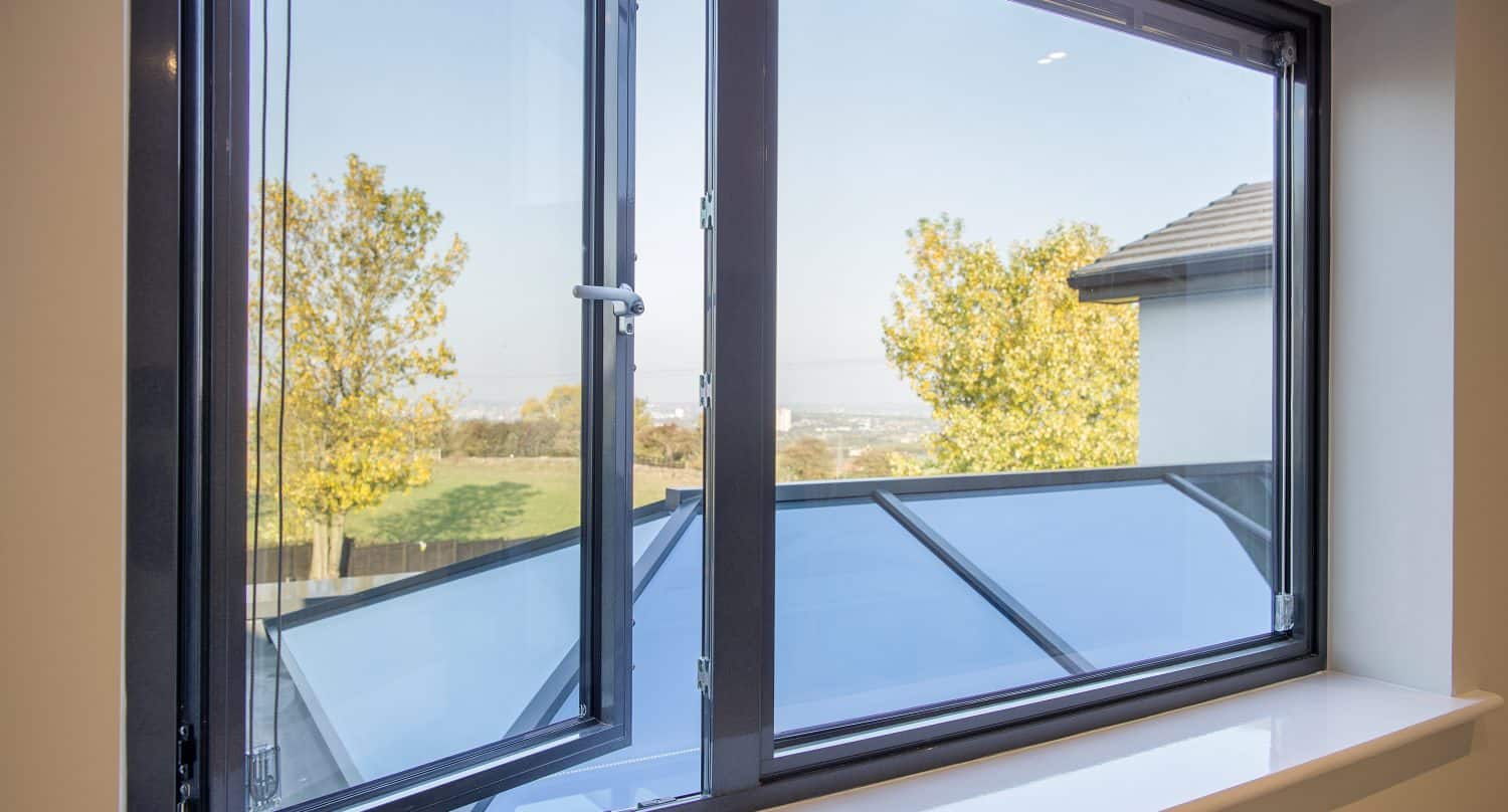 Open window with dark frame, showing a view of trees with autumn leaves, a distant landscape, and part of a roof featuring bespoke glass solutions. The bright daytime scene highlights elegant glass roofing outside.