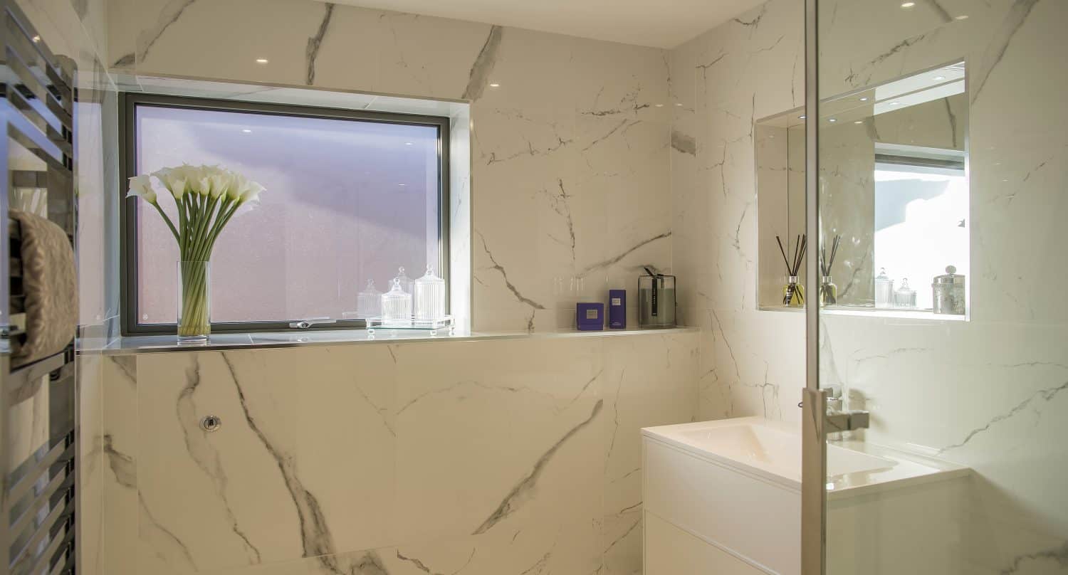Modern bathroom with white marble walls, a bespoke glass shower enclosure, a white sink cabinet, and a window with a vase of calla lilies. The space is brightly lit, minimalistic, and features elegant bespoke glass solutions.
