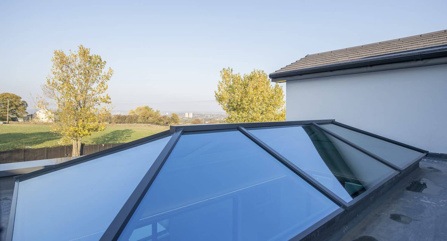 Large glass roofing skylight on a flat rooftop reflects the blue sky, with trees, a grassy area, and a distant cityscape visible in the background—an example of bespoke glass solutions enhancing modern architecture.