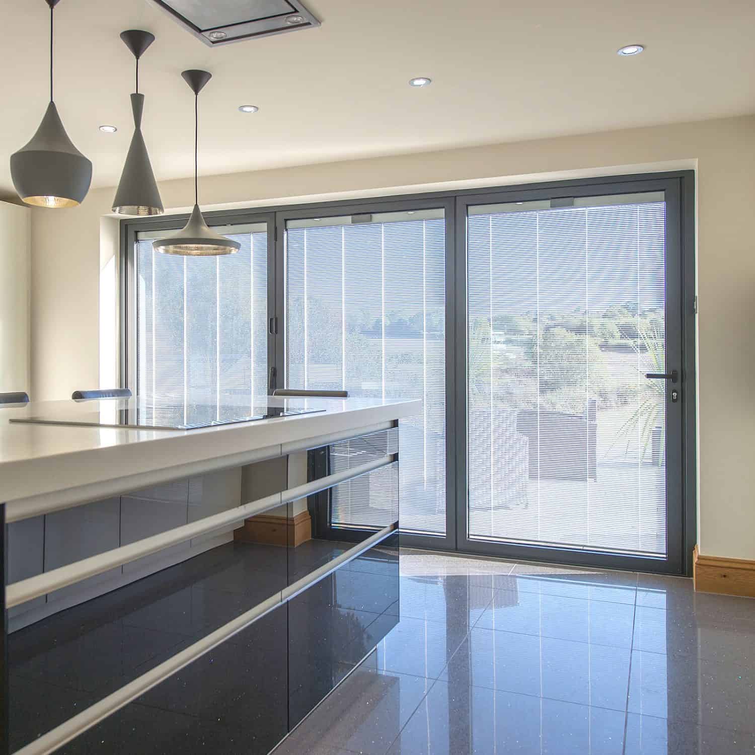 Modern kitchen with glossy black cabinets, a white countertop, and three pendant lights. Large sliding glass doors with sheer blinds let in natural light and feature bespoke glass solutions, offering a stunning view of greenery outside.