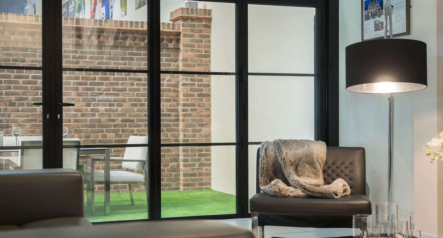 Modern living room with a black leather chair, draped with a fur throw, near a tall floor lamp. Large front doors open to a patio with table and chairs against a brick wall backdrop and green artificial grass flooring.