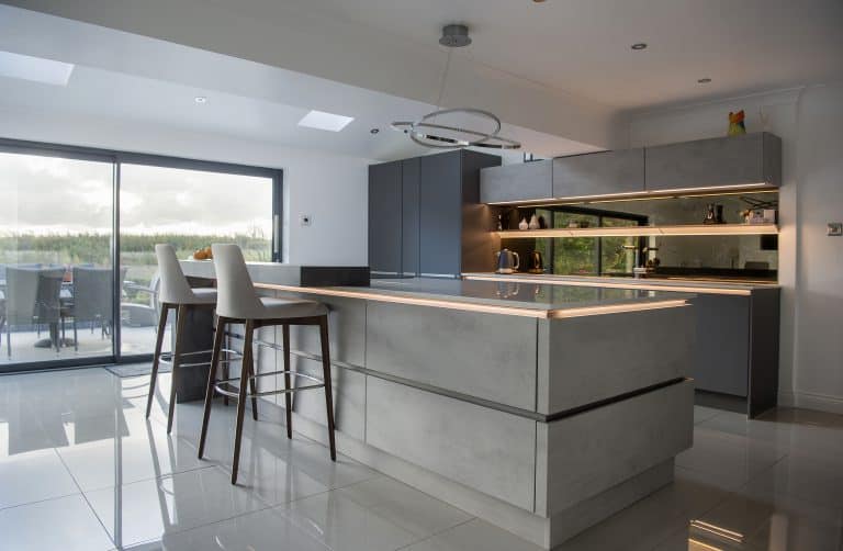 Modern kitchen with sleek grey cabinets, a large island with under-cabinet lighting, two high chairs, reflective tiled floor, built-in appliances, open shelving, and expansive sliding doors leading to an outdoor patio.