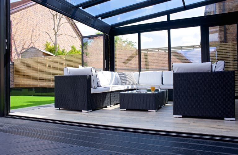 Modern patio with black wicker furniture and white cushions under sleek glass roofing, surrounded by glass walls and sliding doors. A low table holds a glass of orange juice. Wooden fence and grassy area are visible outside.