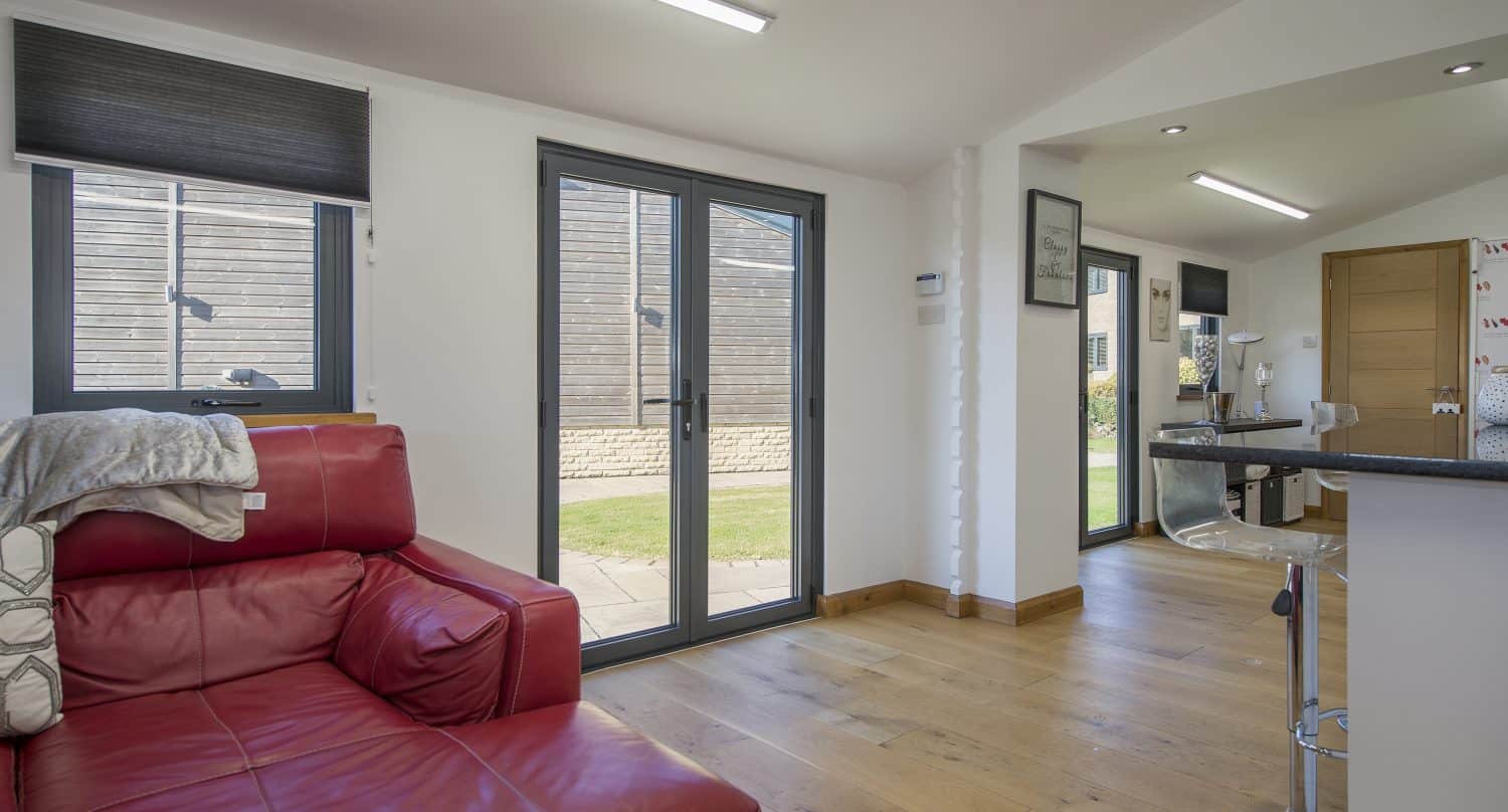 A modern living space with wooden floors, a red leather sofa, and large sliding doors letting in natural light. Bespoke glass solutions enhance the airy feel, while a kitchen area with bar stools and a wooden door is visible in the background.