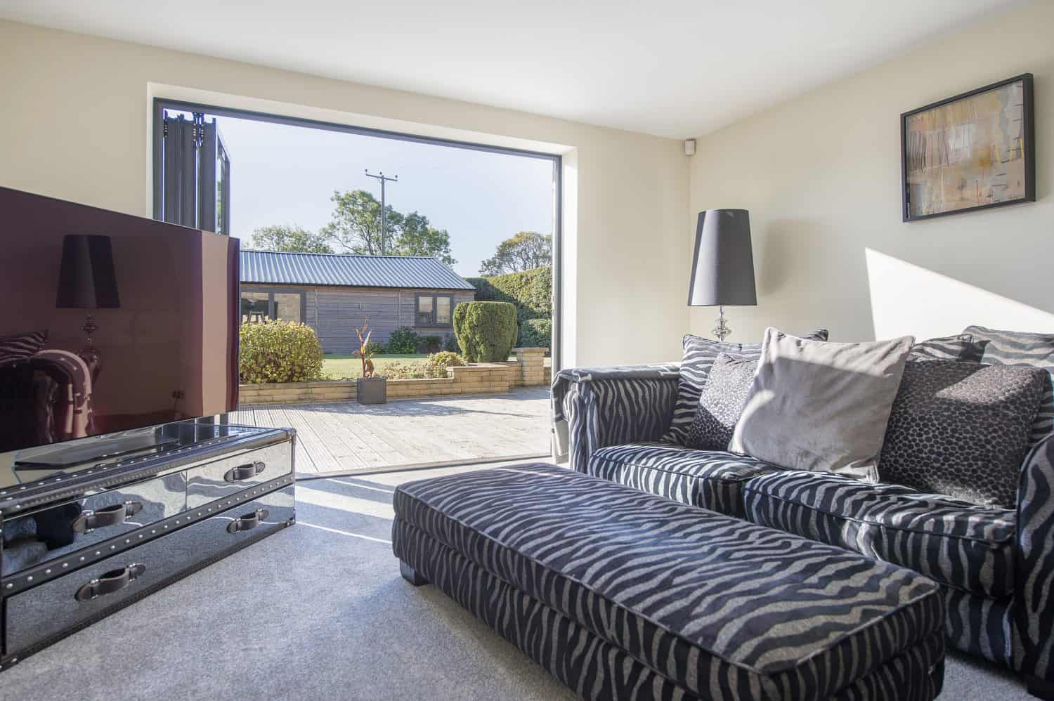 Bright living room with a zebra-patterned sofa and ottoman, a glass TV stand with a large TV, and sliding doors leading to a sunny patio and garden with shrubs and a small building outside.