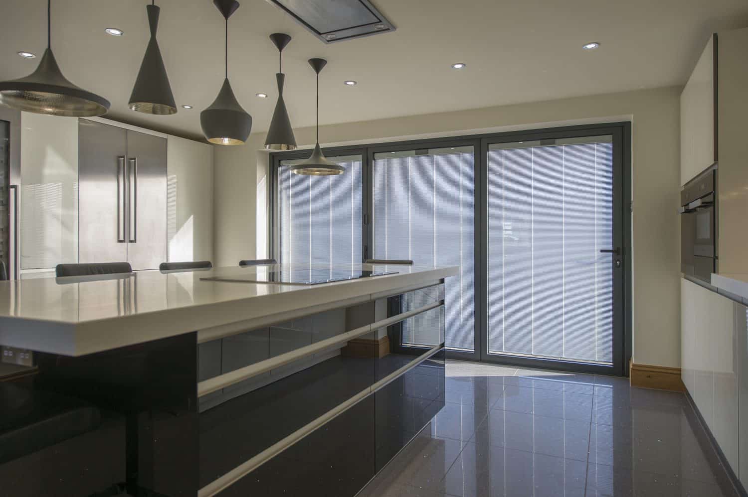 Modern kitchen with glossy black cabinets, a large white island, pendant lights, built-in appliances, and bifold doors letting in natural light. The space appears sleek and minimalistic with bespoke glass solutions enhancing its style.