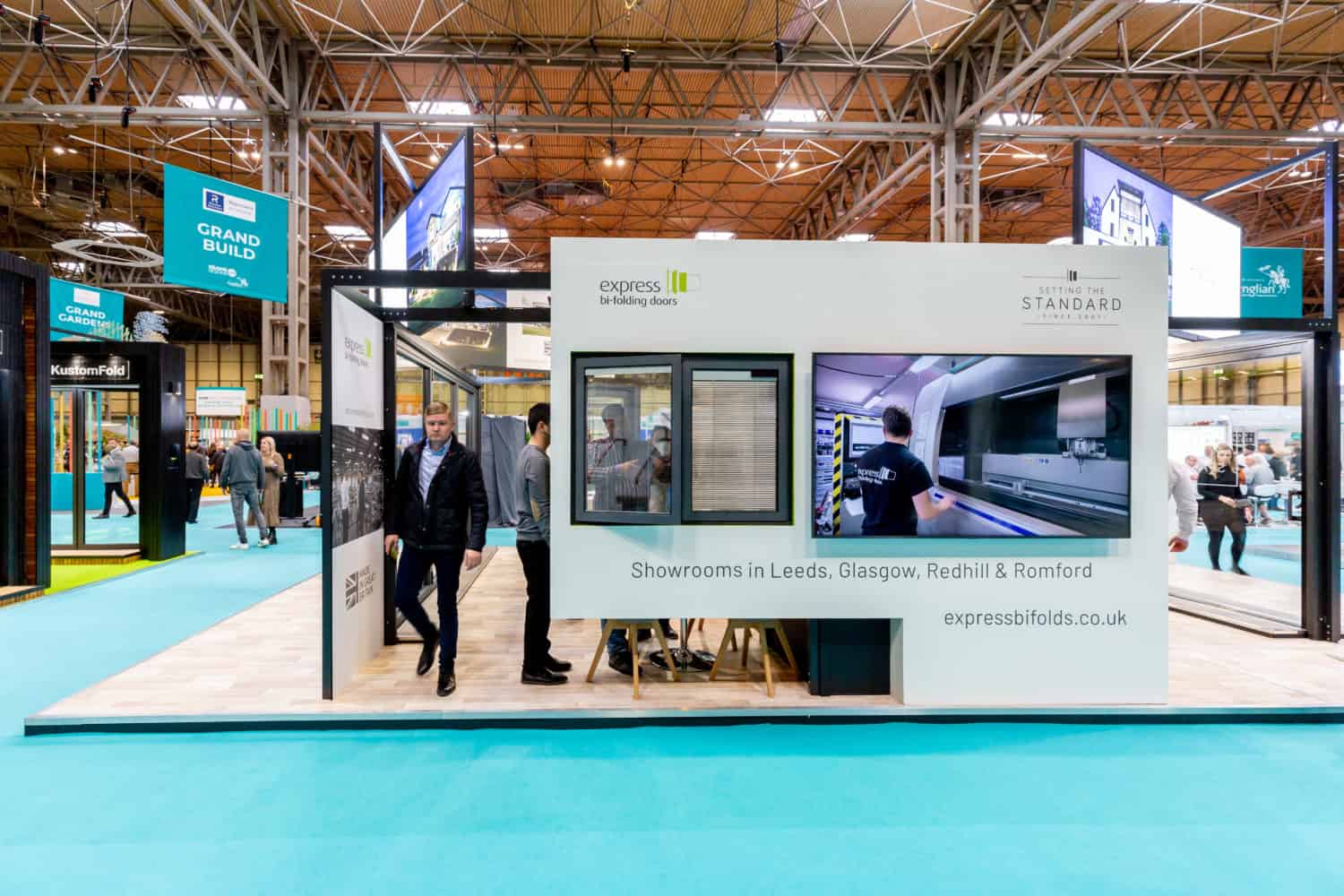 A modern exhibition booth with large video screens, windows, and sliding doors displayed. People walk by and inside the booth featuring bifold doors. Signs promote expressbifolds.co.uk showrooms in a bright, spacious event hall.