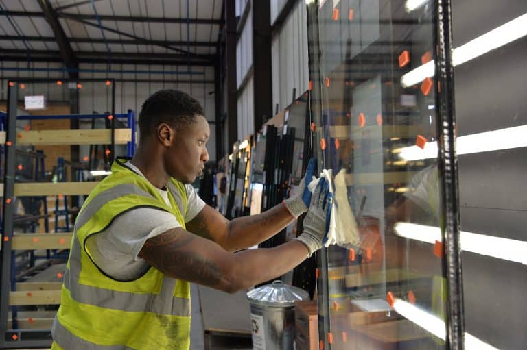 A worker in a high-visibility vest and gloves cleans a large glass pane in an industrial warehouse, surrounded by racks of glass destined for front doors and bespoke glass solutions, illuminated by bright overhead lights.