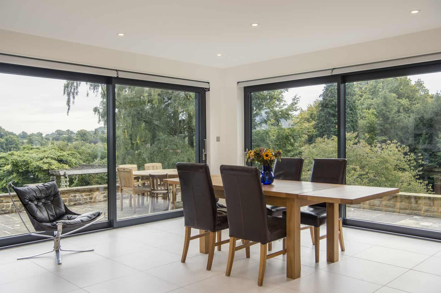 Modern dining room with large glass windows and sliding doors, a wooden table with six chairs, a vase of flowers, a black lounge chair, and a view of the patio and greenery outside.