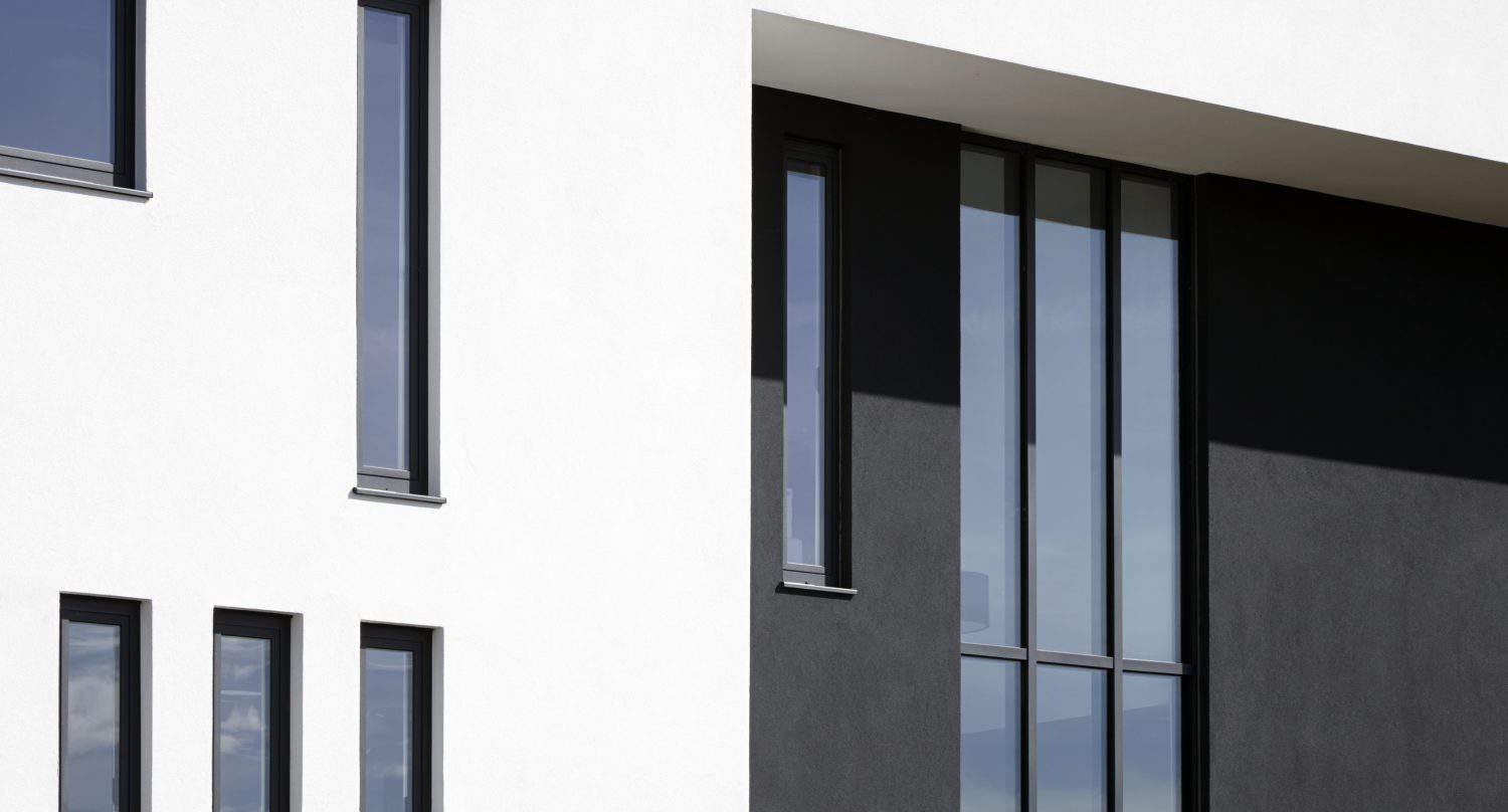 Modern building facade with white and dark gray walls, featuring tall, narrow rectangular windows that reflect the sky. The architectural design emphasizes clean lines and geometric shapes with bespoke glass solutions for a sleek finish.