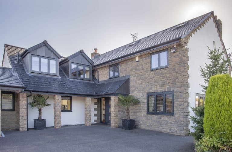 A modern two-story house with stone and white exterior, large windows, sliding doors, a dark sloped roof, and two potted palm trees by the entrance. The driveway is spacious, with the sun visible in the background.