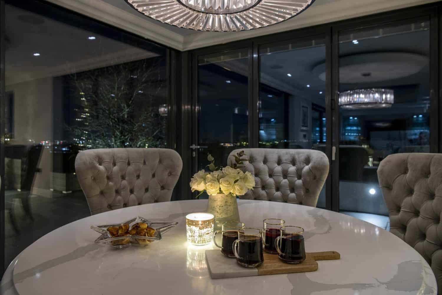 A round marble table with three tufted chairs, four glasses of dark beverage on a wooden board, a lit candle, white flowers, and snacks, set in an elegant dining room with large windows and stylish sliding doors.
