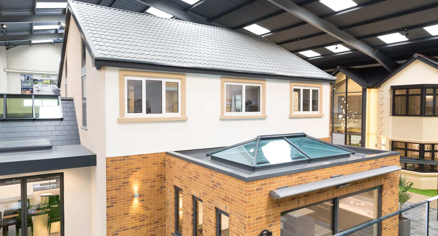 A modern two-story house model is displayed indoors, featuring a brick lower level, cream upper walls, large windows, and a glass roofing extension with bifold doors under a high, skylit ceiling.