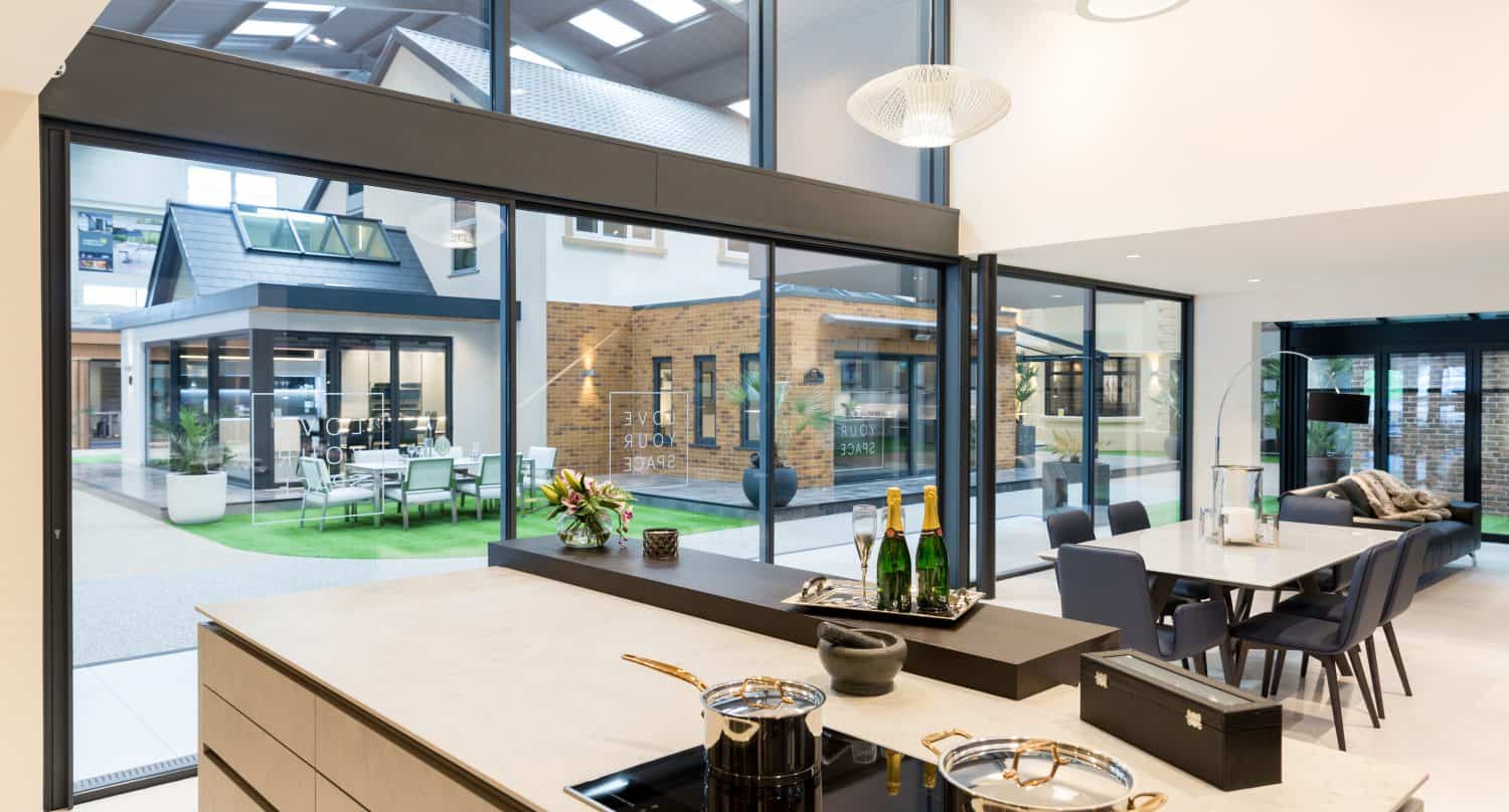 Modern kitchen and dining area with large windows and sliding doors overlooking a bright courtyard and neighboring buildings. The space features a sleek island, induction cooktop with pots, and elegant table settings.
